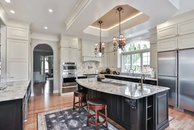 Large white kitchen with a dark curved island, granite counters, tray ceiling and twin chandeliers